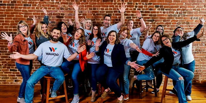 Group of coworkers in matching WAHOO t-shirts posing together with arms raised in a brick-walled room, smiling and cheerful.